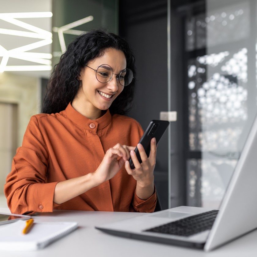 Woman tapping on phone with laptop in front of her